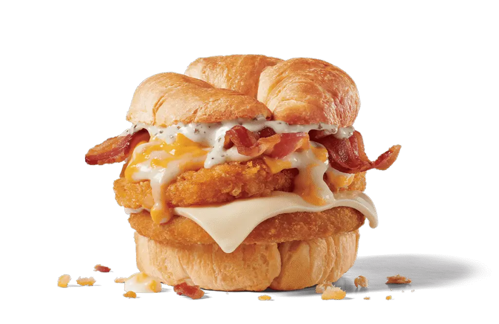 A mouthwatering Chick-N-Tater Melt sandwich from Jack in the Box. The sandwich features crispy chicken layered with melted cheese, golden hash browns, savory bacon, and a rich, creamy white sauce, all nestled between two buttery croissant buns. Crumbs are scattered around, showcasing its flaky texture.