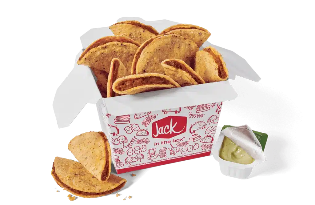 A box of classic tiny tacos from Jack in the Box, featuring small, crispy taco shells filled with seasoned beef. The tacos are presented in a branded red and white box with a design of playful illustrations. A container of green avocado lime sauce, partially opened, is placed beside the box, and a few tacos are scattered in front.