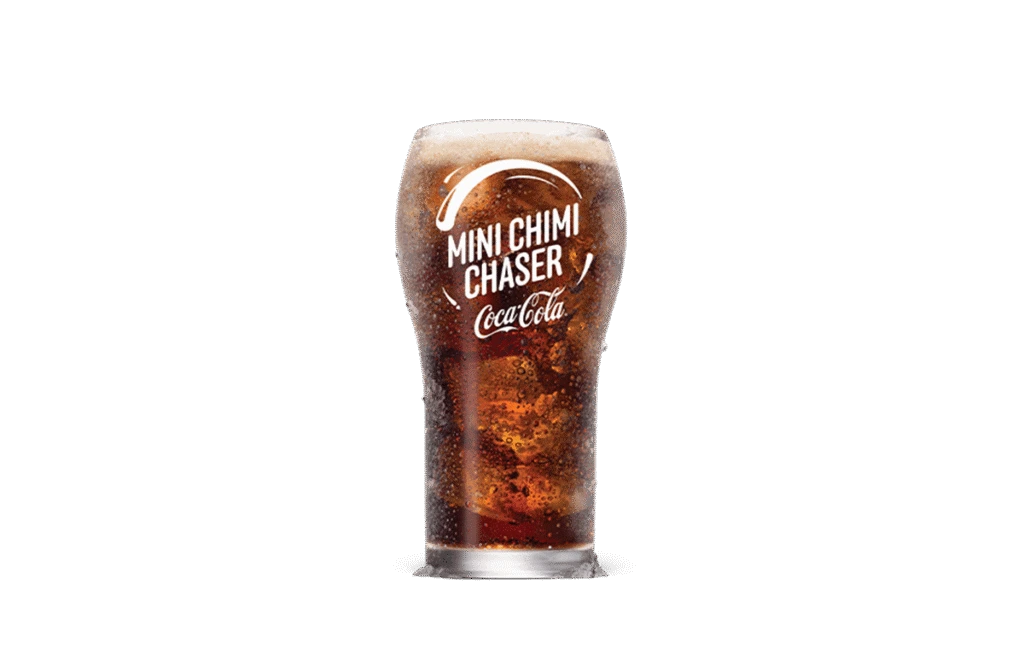 A glass of Coca-Cola® Mini Chimi Chaser with visible carbonation and foam on top. The glass features the Coca-Cola® logo and "Mini Chimi Chaser" text in white, emphasizing its refreshing and fizzy appearance. The dark background enhances the drink's vibrant, bubbly texture.