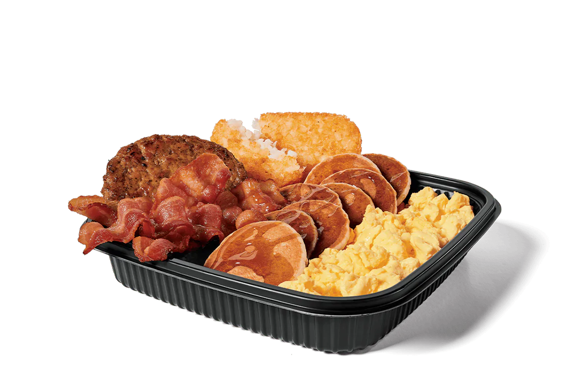 Image of a jumbo breakfast platter featuring crispy bacon strips, two sausage patties, fluffy scrambled eggs, golden hash browns, and mini pancakes drizzled with syrup, all served in a black plastic tray.