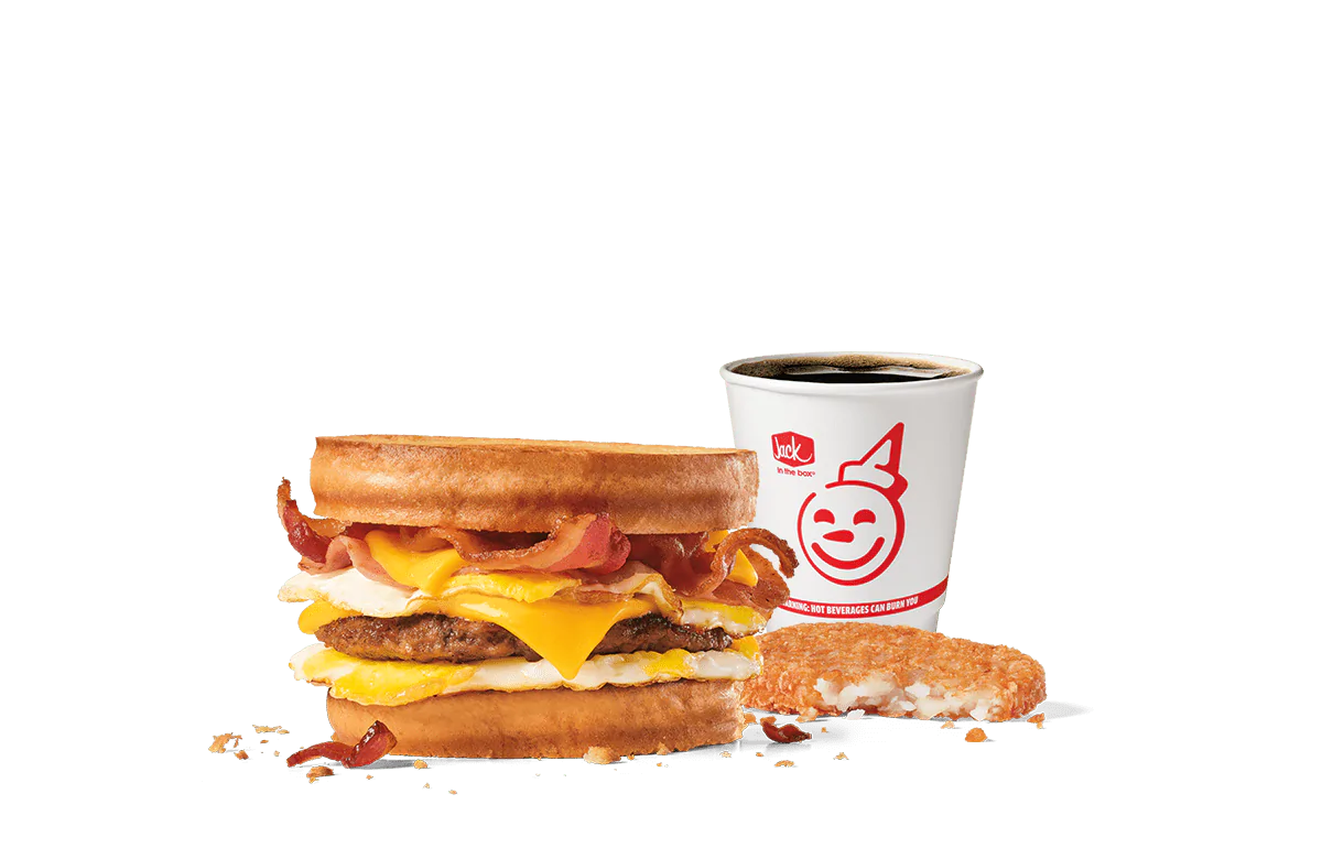 Image of the Loaded Breakfast Sandwich Combo from Jack in the Box, featuring a hearty sandwich with crispy bacon, a sausage patty, egg, and cheese between toasted sourdough bread. Served with a hash brown and a cup of coffee, all presented against a transparent background with the cheerful Jack in the Box logo.