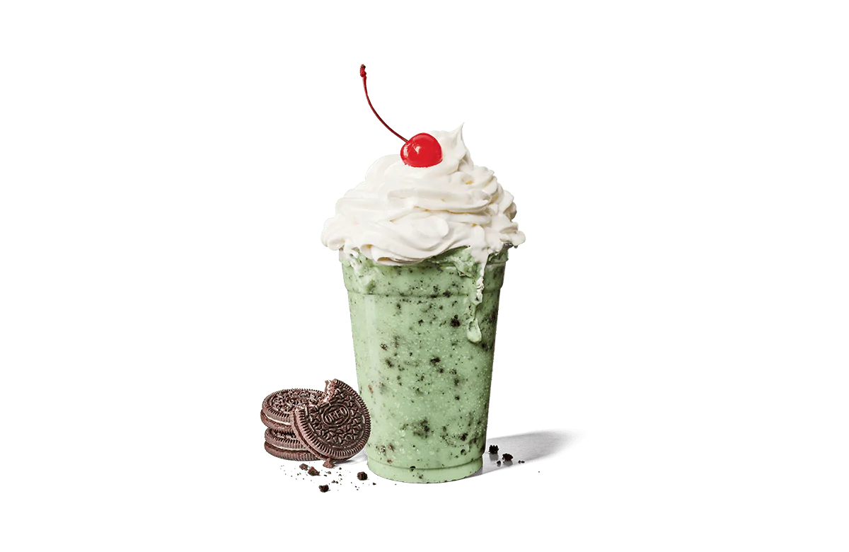 A refreshing Oreo® cookie mint shake in a clear cup, topped with a generous amount of whipped cream and a single cherry. The shake has a vibrant mint green color and is speckled with crushed Oreo® cookie pieces. Several whole Oreo® cookies lie beside the cup transparent background, some crumbled, adding to the dessert's indulgent appeal.