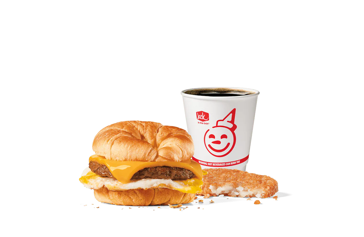 Image of the Sausage Croissant Combo from Jack in the Box, featuring a breakfast sandwich with a savory sausage patty, fried egg, and melted cheese nestled inside a flaky croissant. Accompanied by a hash brown and a cup of coffee, all set against a transparent background with the iconic smiling Jack logo.