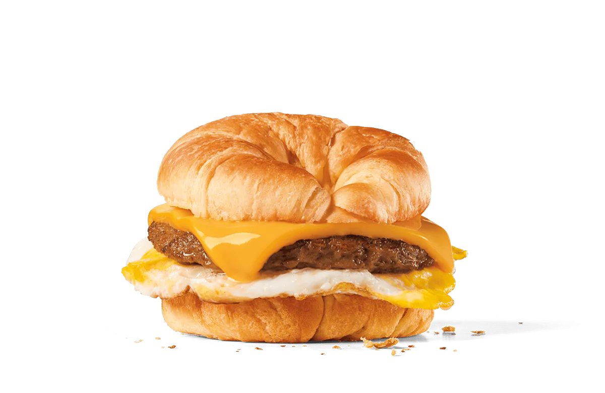 A hearty breakfast sandwich featuring a flaky croissant split and filled with a savory sausage patty, a slice of melted American cheese, and a fried egg, set against a transparent background.
