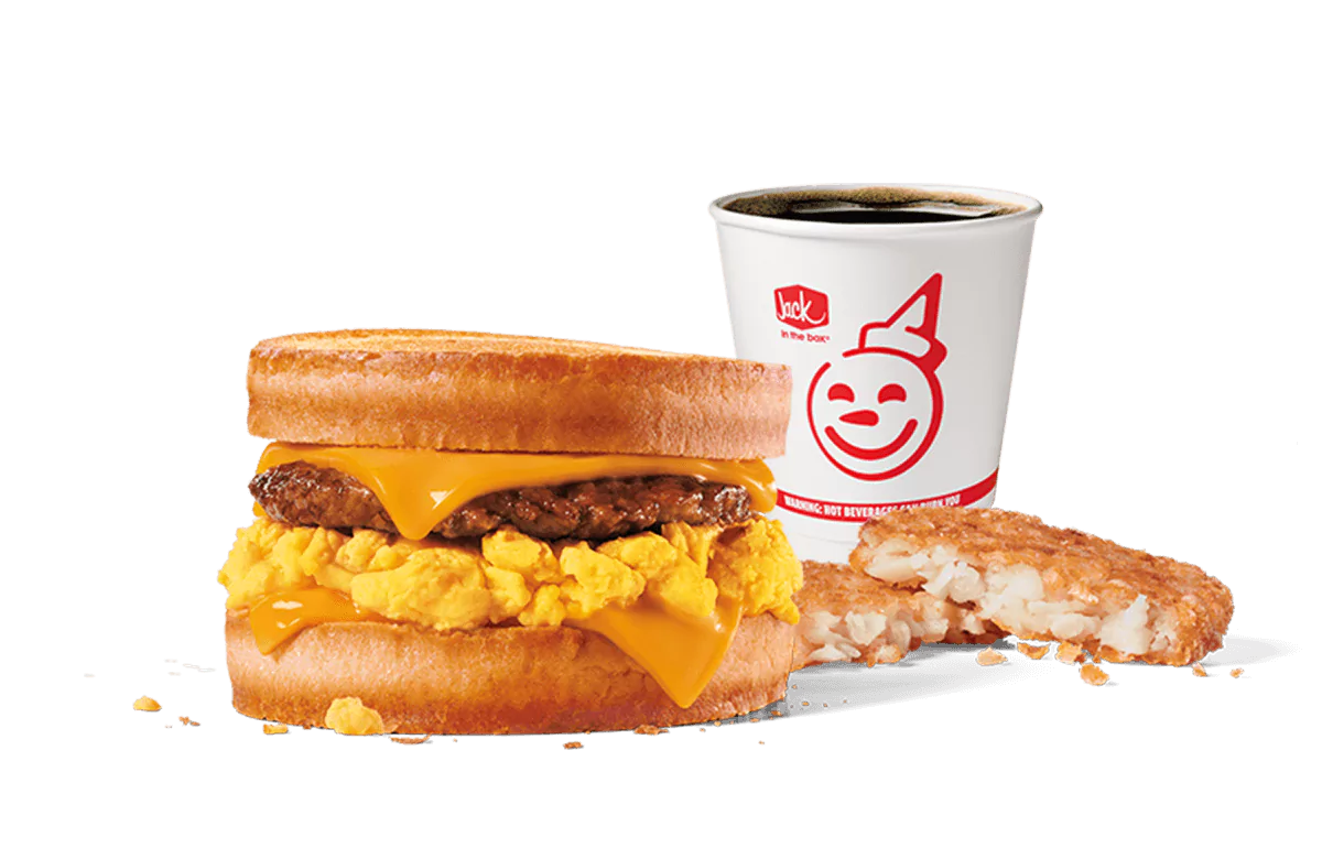 Image of Jack in the Box's Sausage, Egg & Cheese Scrambler Combo, featuring a breakfast sandwich with a sausage patty, scrambled eggs, and melted cheese on a muffin, alongside a hash brown and a coffee cup with the iconic smiling face logo. The backdrop is transparent, focusing attention on the rich colors and textures of the meal components.