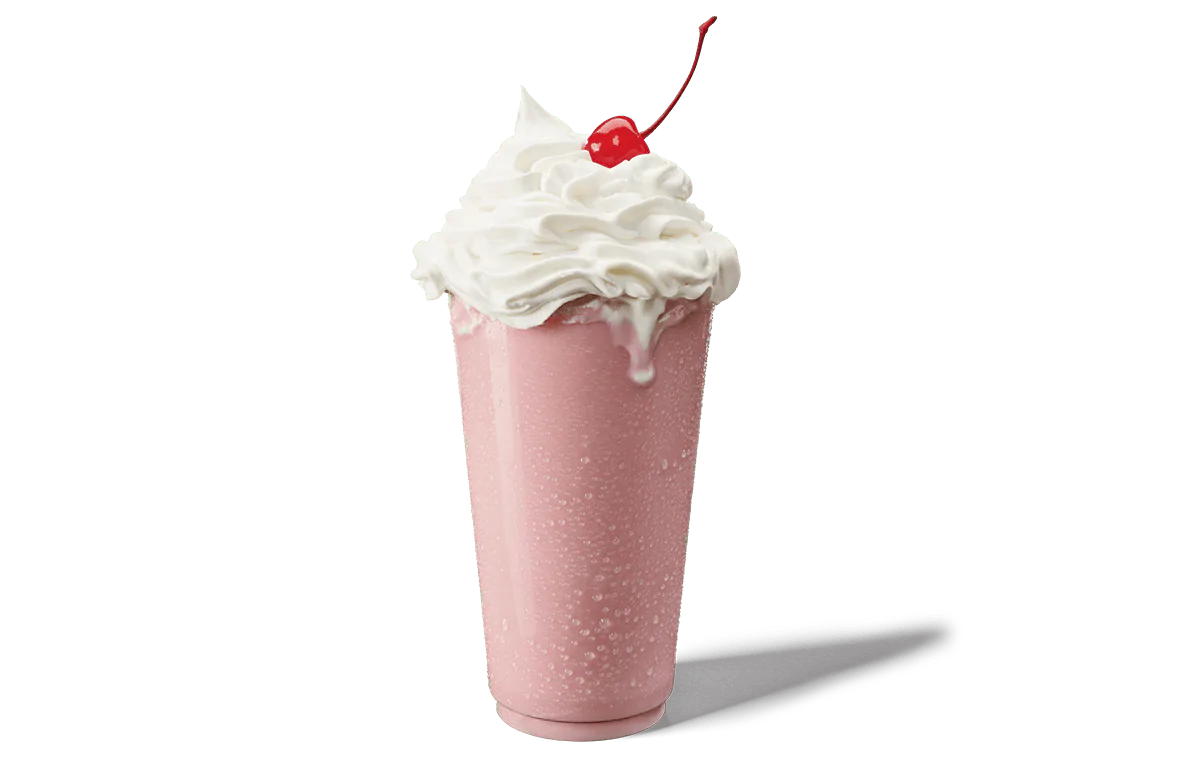 A strawberry shake in a tall glass, topped with an abundant swirl of whipped cream and a bright red cherry. The shake is a soft pink color, with visible condensation on the glass, suggesting a cold and refreshing beverage. The whipped cream overflows slightly, adding to the indulgent look of this classic dessert.