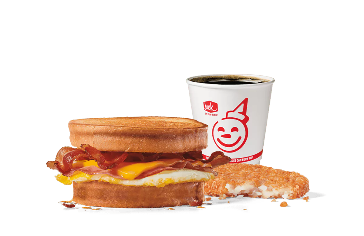 Image of the Supreme Sourdough Breakfast Sandwich Combo from Jack in the Box, featuring crispy bacon, a fried egg, and melty cheese between toasted sourdough slices. Accompanied by a hash brown and a cup of hot coffee, all set against a transparent background with the iconic smiling Jack logo.