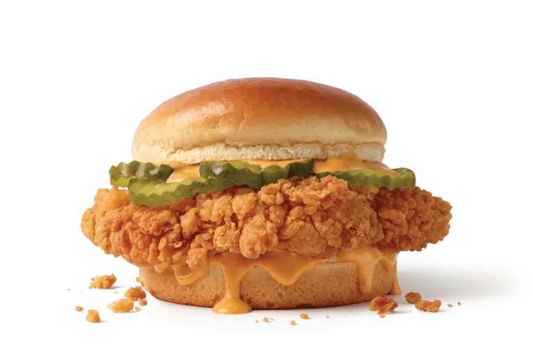 A sumptuous crispy chicken sandwich loaded with thick breaded chicken, topped with tangy pickles and drizzled with spicy sauce, nestled between a soft, shiny bun, with crumbs scattered around on a transparent background.