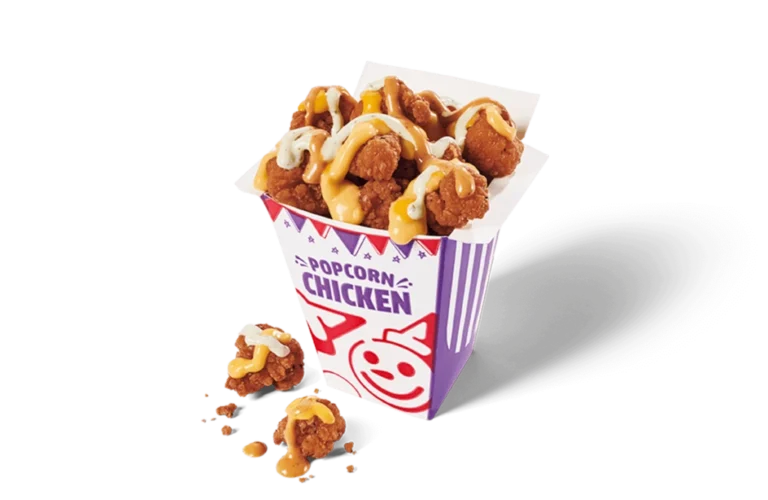 A vibrant carton overflowing with spicy popcorn chicken, generously topped with swirls of yellow and white sauces. The nuggets are golden-brown and crispy, some spilling out onto the transpparent background, with crumbs visible around the carton.