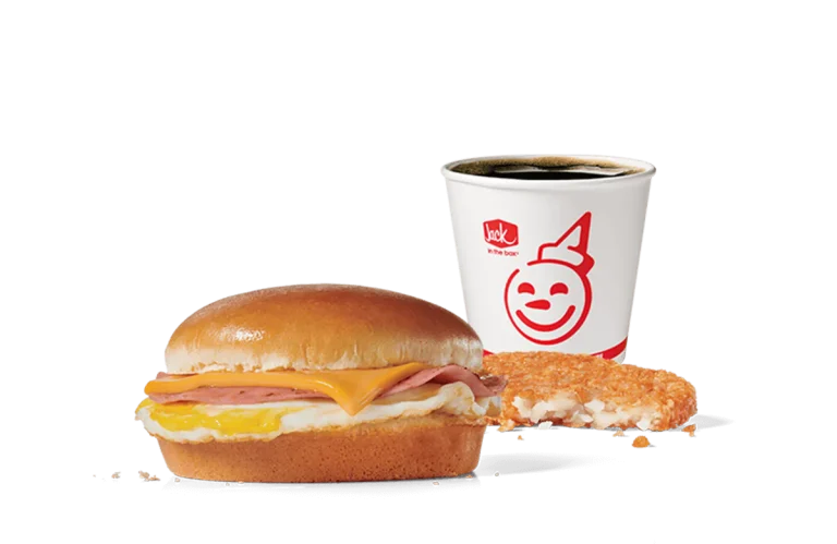 A Breakfast Jack® Combo from Jack in the Box featuring a classic sandwich with ham, egg, and American cheese on a toasted bun, alongside a crispy hash brown and a cup of hot coffee. The set is displayed against a transparent background.