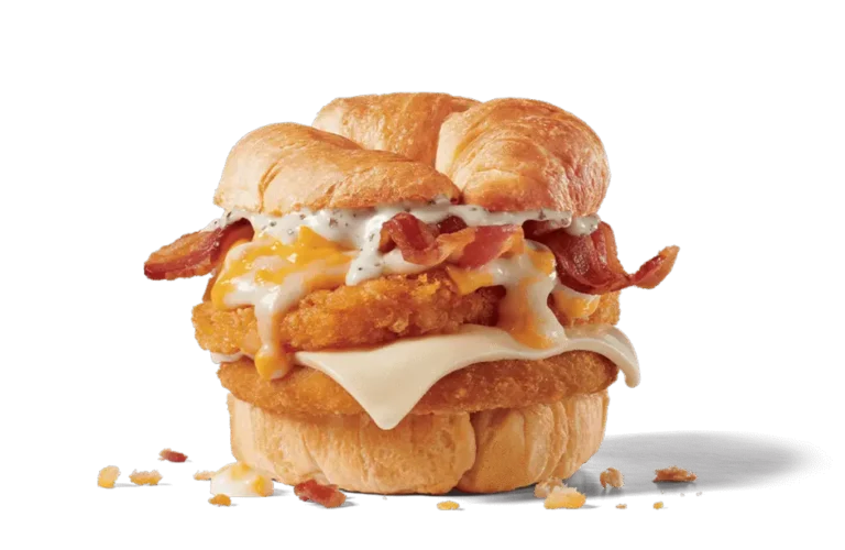 A mouthwatering Chick-N-Tater Melt sandwich from Jack in the Box. The sandwich features crispy chicken layered with melted cheese, golden hash browns, savory bacon, and a rich, creamy white sauce, all nestled between two buttery croissant buns. Crumbs are scattered around, showcasing its flaky texture.