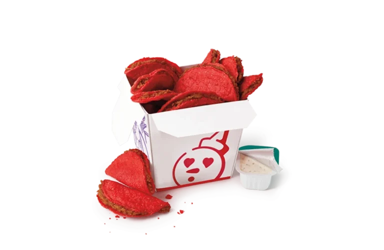 A box of classic spicy tiny tacos from Jack in the Box, featuring mini tacos with a bright red, spicy coating. The tacos are served in a white box with red and purple designs, including a heart-eyed character illustration. A few tacos are displayed outside the box alongside a cup of creamy dipping sauce.