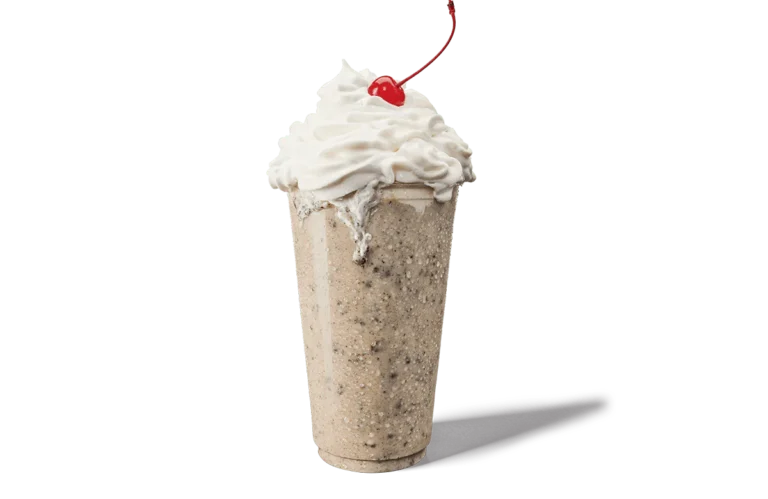 A tall, creamy Oreo® shake overflowing with whipped cream, topped with a single bright red cherry. The shake is speckled with pieces of Oreo® cookies throughout, giving it a textured look against transparent background. The whipped cream is generously swirled on top, creating an appealing and indulgent dessert.