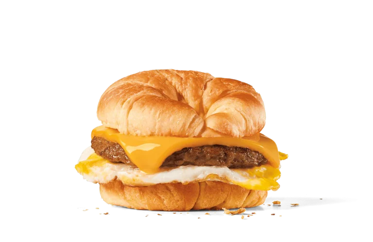 A hearty breakfast sandwich featuring a flaky croissant split and filled with a savory sausage patty, a slice of melted American cheese, and a fried egg, set against a transparent background.