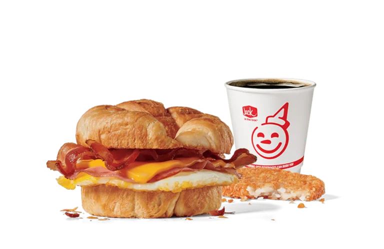 Image of the Supreme Croissant Combo from Jack in the Box, featuring a breakfast sandwich with crispy bacon, a fried egg, and melty cheese inside a flaky croissant. Accompanied by a hash brown and a cup of coffee, all set against transparent background with the iconic Jack in the Box logo.